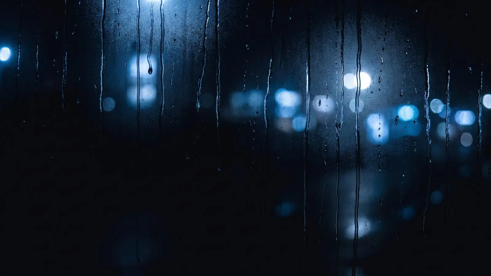 Rain on a dark window at night with out-of-focus blue city lights behind.