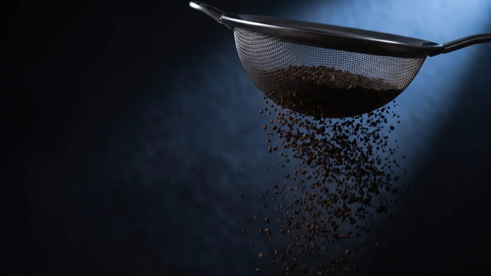 Dark grains falling through a fine sieve, lit from one side.